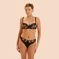 A woman modeling a black lace bra and matching lace underwear against a solid beige studio background.