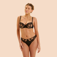 A woman modeling a black lace lingerie set against a solid beige studio background. The set includes a sheer floral lace underwire bra and matching lace-trimmed briefs.