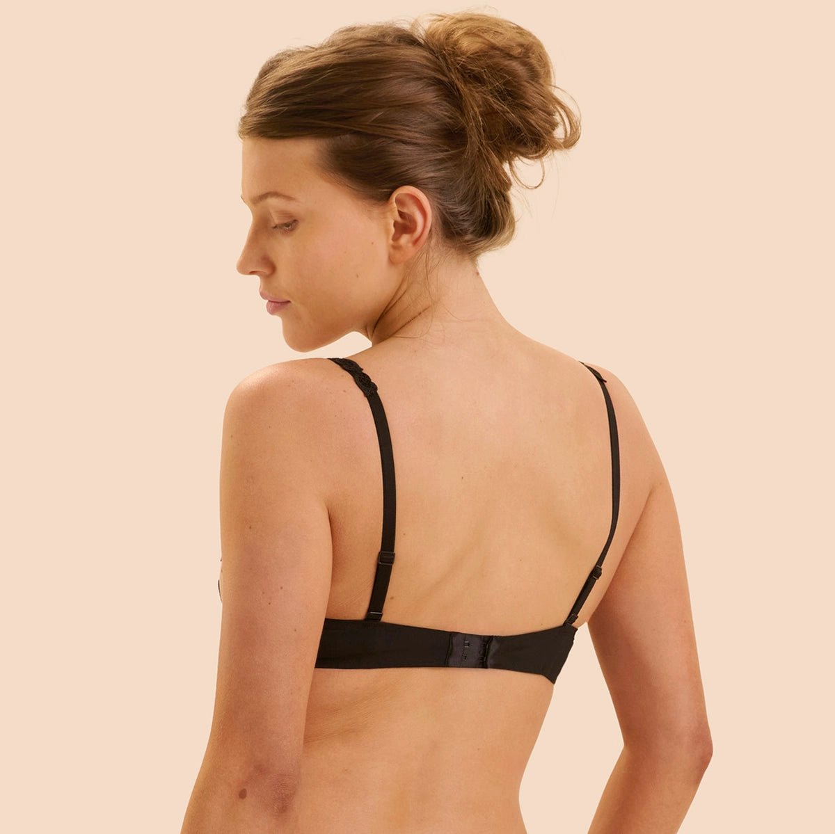 A woman shown from the back wearing a black underwire bra with thin adjustable shoulder straps and a center back strap, against a solid light beige background.