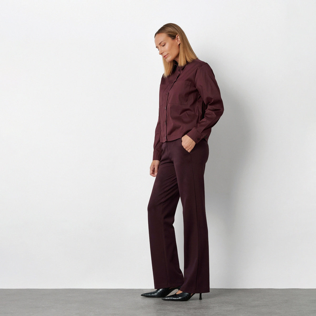 A woman stands in profile wearing a matching burgundy button-up shirt and wide-leg trousers set, paired with black pointed-toe flats, against a plain light gray studio wall.