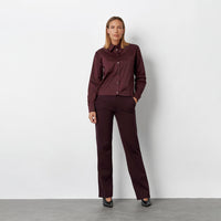 A woman stands against a light gray studio wall wearing a matching burgundy button-up shirt and trousers set with black pointed shoes.