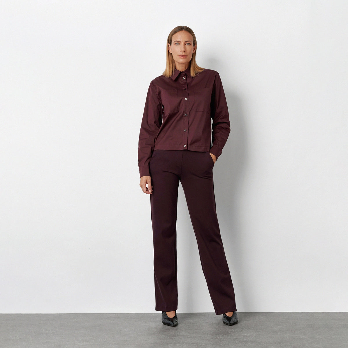 A woman stands against a light gray studio wall wearing a matching burgundy button-up shirt and trousers set with black pointed shoes.