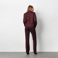 A back view of a woman  wearing a matching burgundy button-up shirt and wide-leg trousers set, paired with black pointed-toe flats, against a plain light grey studio wall.