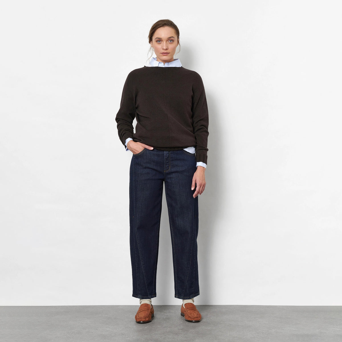 A woman stands against a light grey studio wall wearing a dark brown knit sweater, dark blue barrel jeans, brown suede loafers, and a white neck scarf.