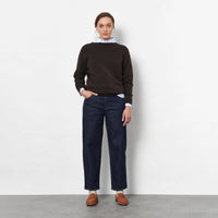 A woman stands against a light grey studio wall wearing a dark brown knit sweater, dark blue barrel jeans, brown suede loafers, and a white neck scarf.