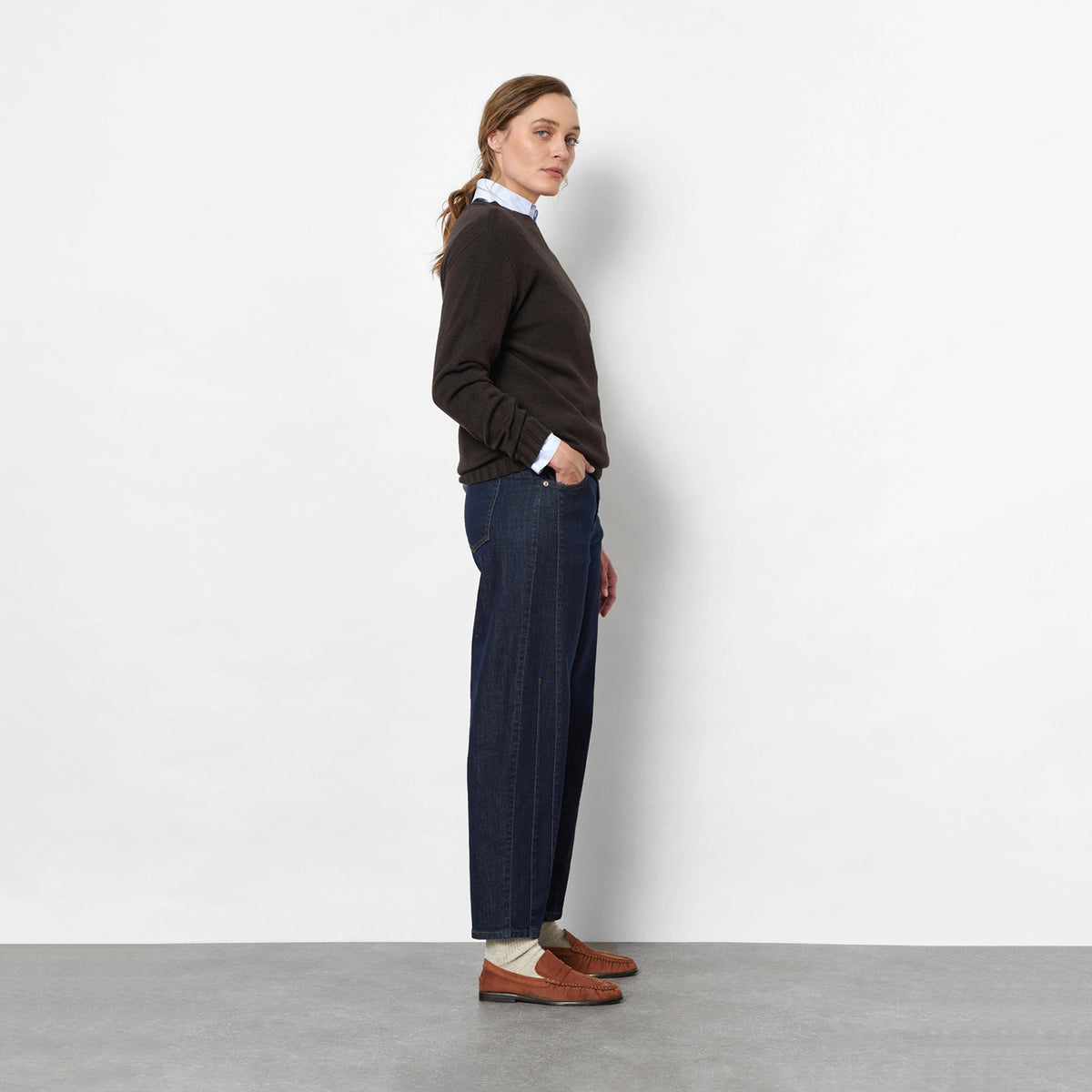 Side view of a woman against a light grey studio wall wearing a dark brown knit sweater, dark blue barrel jeans, brown suede loafers, and a white neck scarf.