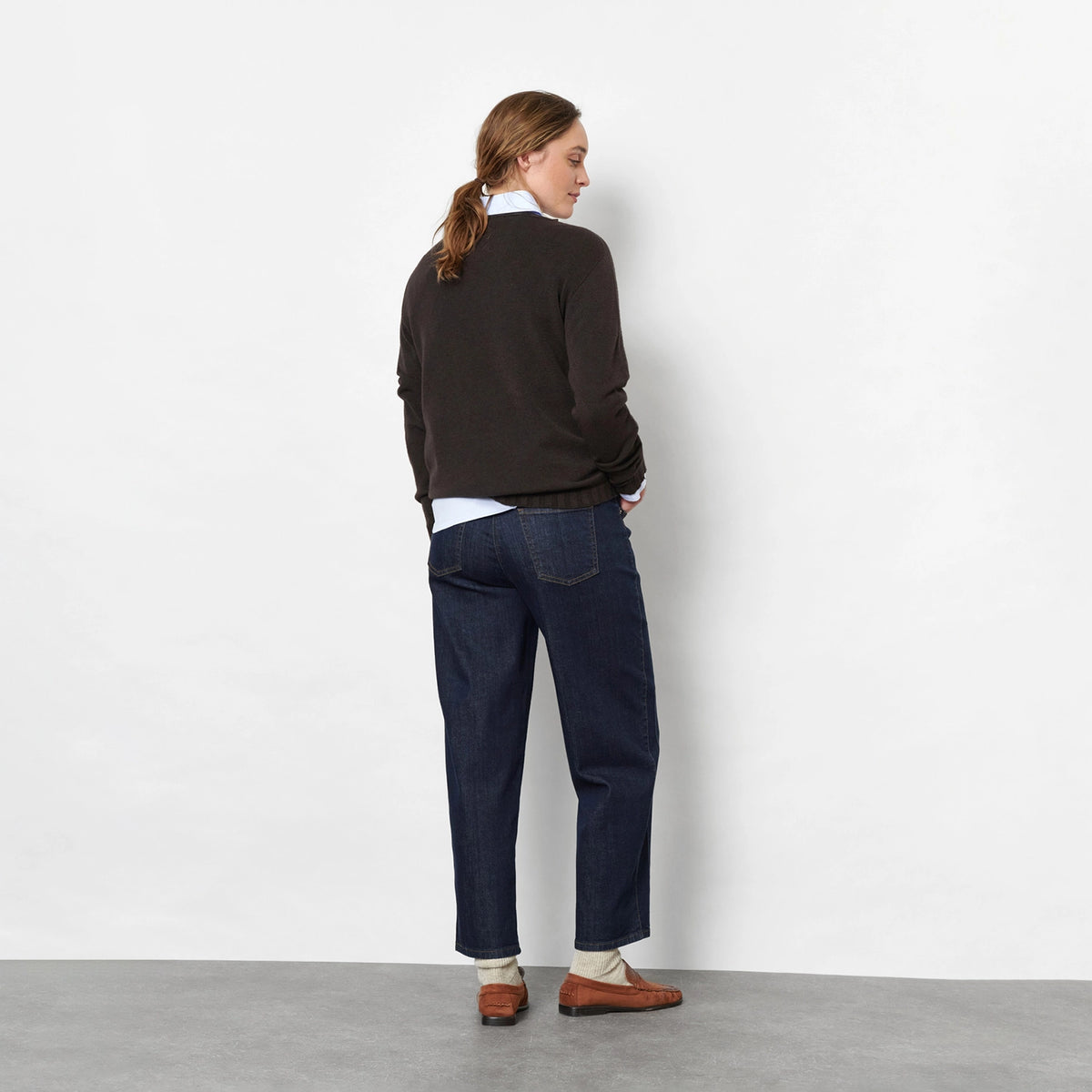 Back view of a woman against a light grey studio wall wearing a dark brown knit sweater, dark blue barrel jeans, brown suede loafers, and a white neck scarf.