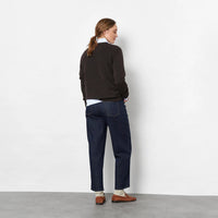 Back view of a woman against a light grey studio wall wearing a dark brown knit sweater, dark blue barrel jeans, brown suede loafers, and a white neck scarf.