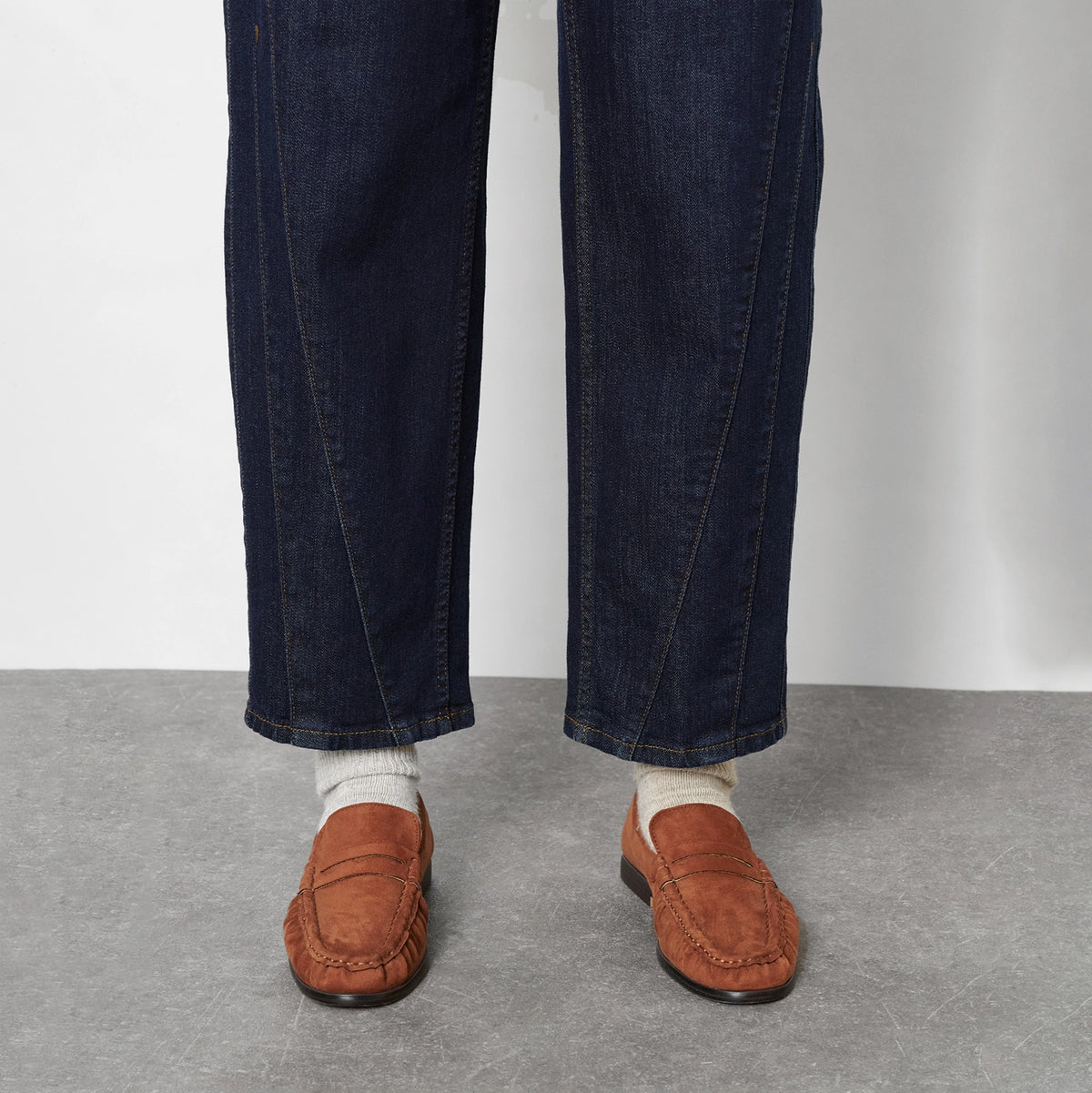 Crop from the knee down of a person wearing dark blue denim jeans, cream knit socks, and brown leather penny loafers, against a light grey wall and concrete floor.