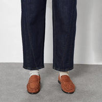 Crop from the knee down of a person wearing dark blue denim jeans, cream knit socks, and brown leather penny loafers, against a light grey wall and concrete floor.