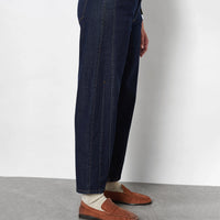 A person wearing dark blue denim jeans, cream knit socks, and brown leather penny loafers, shown from the waist down against a light gray wall and concrete floor.
