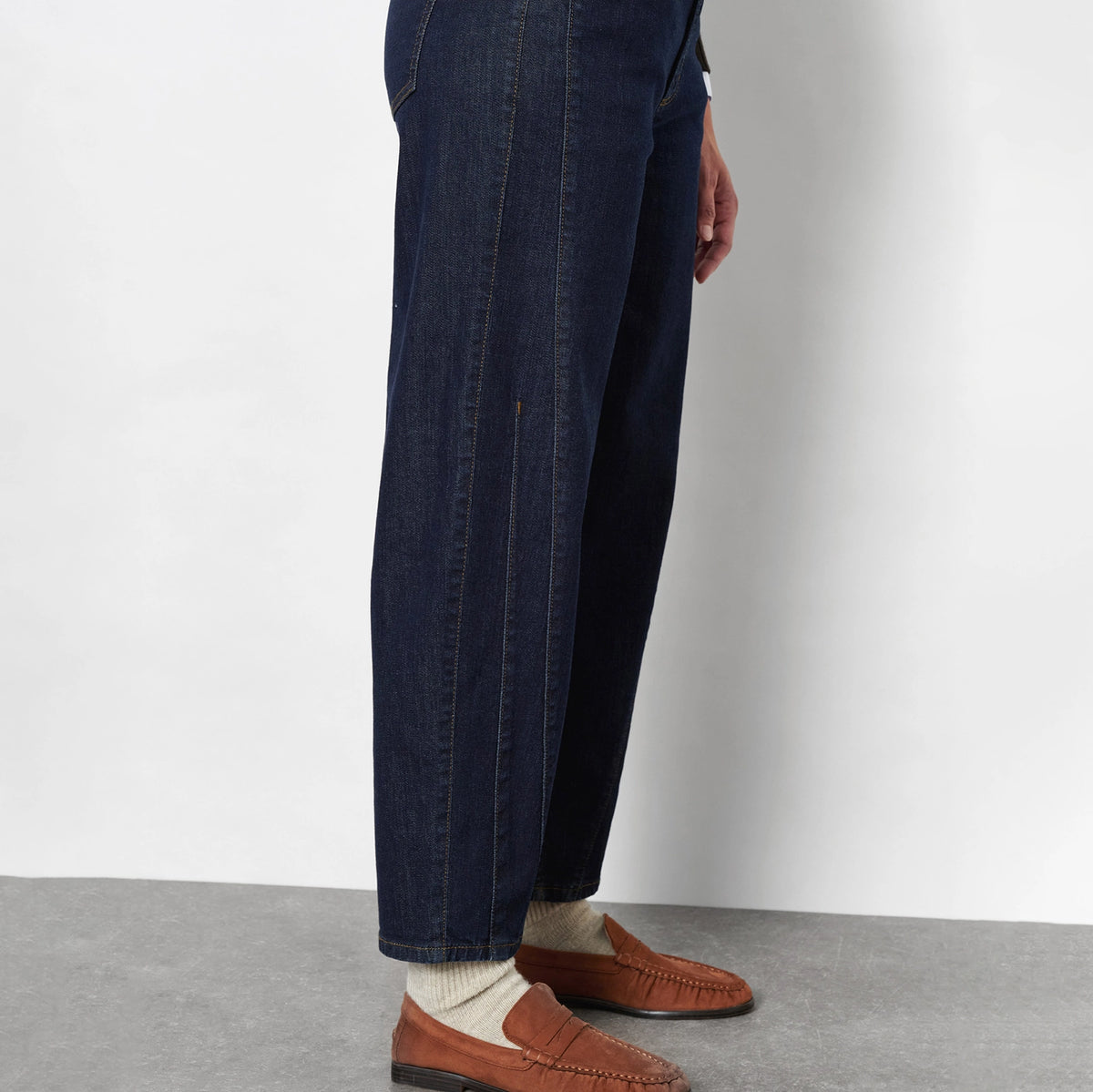 A person wearing dark blue denim jeans, cream knit socks, and brown leather penny loafers, shown from the waist down against a light gray wall and concrete floor.