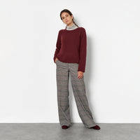 A woman against a plain white wall, wearing a burgundy long-sleeve top, a light gray and white striped turtleneck, wide-leg gray plaid trousers, and black shoes.