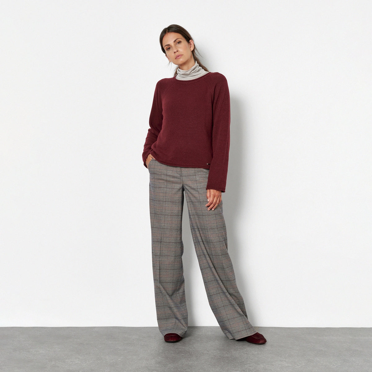 A woman against a plain white wall, wearing a burgundy long-sleeve top, a light gray and white striped turtleneck, wide-leg gray plaid trousers, and black shoes.