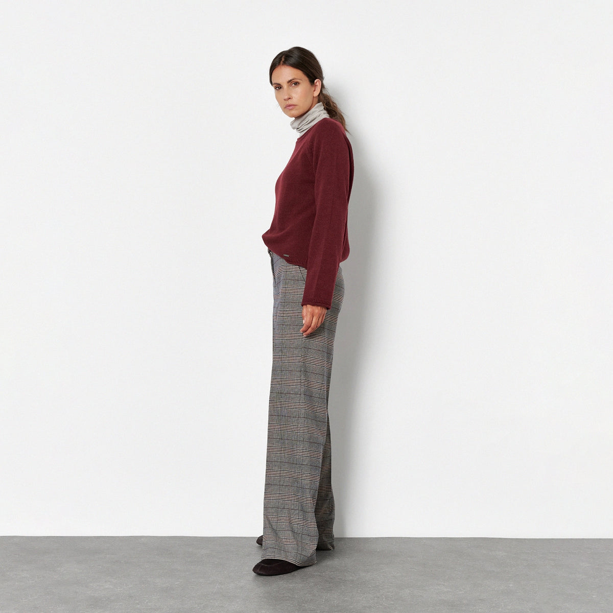 A woman stands in profile against a plain white wall, wearing a burgundy long-sleeve top, a light gray and white striped turtleneck, wide-leg gray plaid trousers, and black shoes.