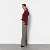 A woman stands in profile against a plain white wall, wearing a burgundy long-sleeve top, a light gray and white striped turtleneck, wide-leg gray plaid trousers, and black shoes.