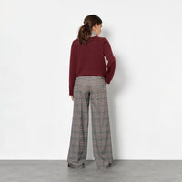 Back view of a woman against a plain white wall, wearing a burgundy long-sleeve top, a light gray and white striped turtleneck and wide-leg gray plaid trousers.