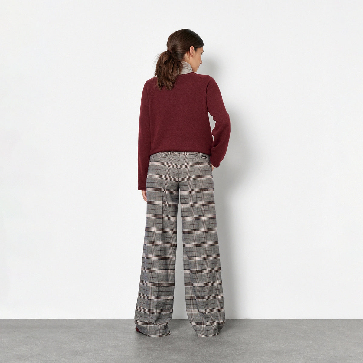 Back view of a woman against a plain white wall, wearing a burgundy long-sleeve top, a light gray and white striped turtleneck and wide-leg gray plaid trousers.