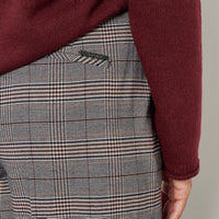 Close-up of a person wearing plaid trousers with a burgundy knit top tucked into the waistband. The trousers feature a fine check pattern in gray, white, and thin red lines, with a visible metal logo on the back pocket.