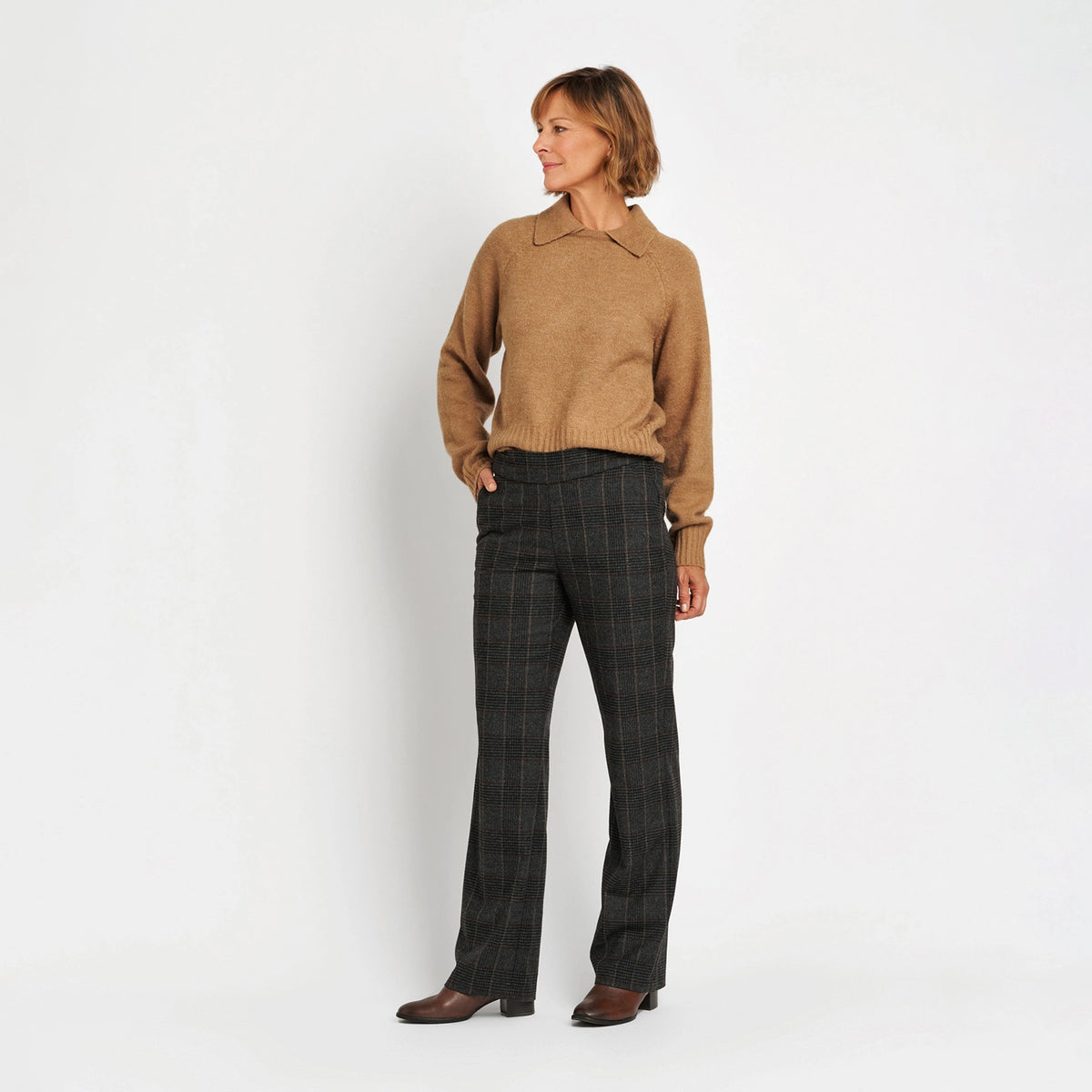A woman stands in profile wearing a camel knit polo-style sweater and dark charcoal plaid trousers with brown shoes, photographed against a clean light gray studio background.