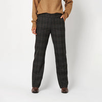 A person wearing dark charcoal gray plaid trousers, a tan knit sweater, and brown shoes, standing against a light gray studio background.