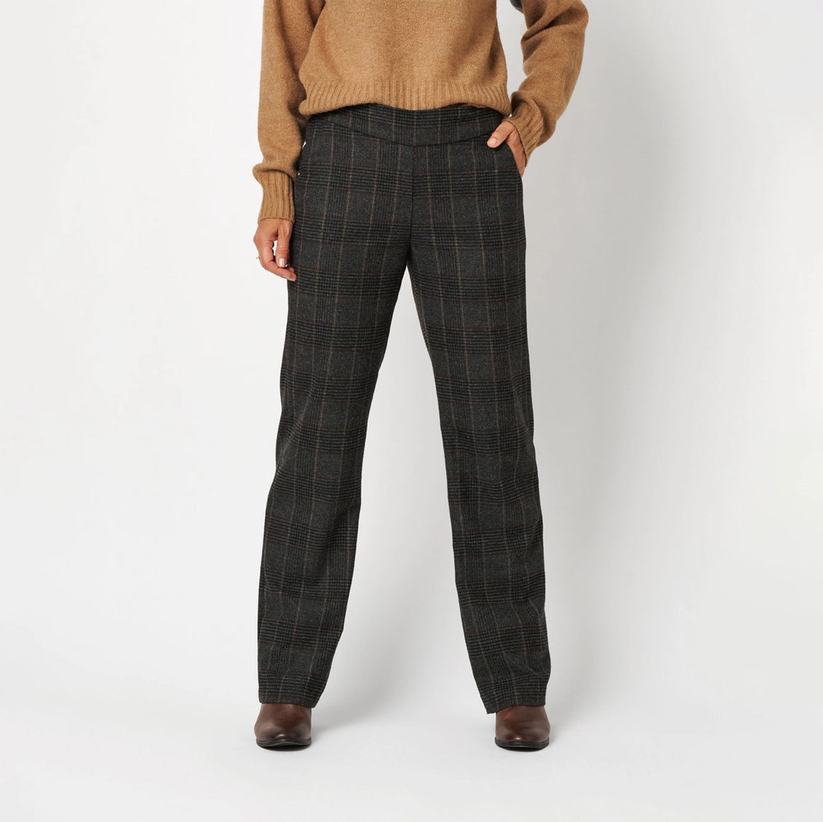 A person wearing dark charcoal gray plaid trousers, a tan knit sweater, and brown shoes, standing against a light gray studio background.