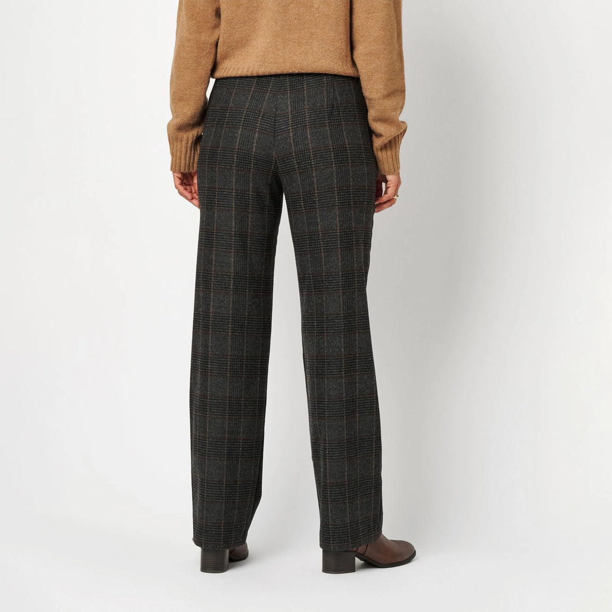 A person shown from the back wearing dark charcoal gray plaid trousers, a tan sweater, and brown shoes against a light gray studio background.