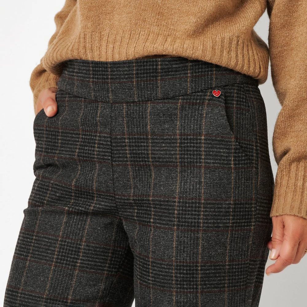 Close-up of a person wearing dark charcoal plaid trousers with a small red heart logo on the pocket, paired with a tan ribbed knit sweater.