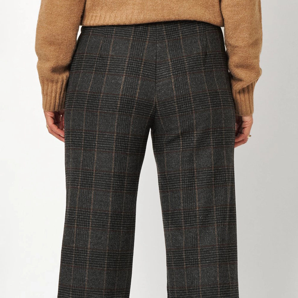Back view of a person wearing dark charcoal gray plaid trousers with a tan knit sweater, shown against a light gray studio background.