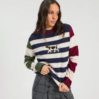 A woman wearing a striped knit sweater with navy, cream, burgundy, and green horizontal bands, featuring a dog graphic on the chest, paired with gray plaid trousers against a light gray studio background.