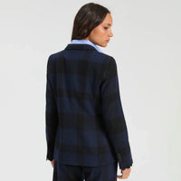 A woman shown from the back wearing a navy and black plaid blazer over a light blue collared shirt, photographed against a plain light gray studio background.