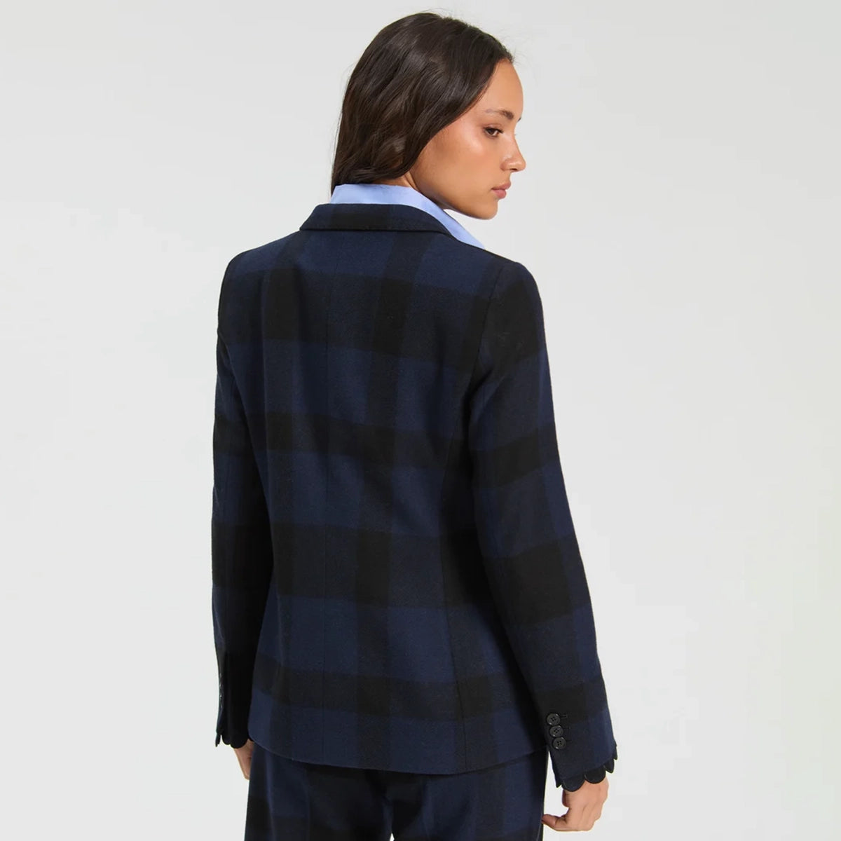A woman shown from the back wearing a navy and black plaid blazer over a light blue collared shirt, photographed against a plain light gray studio background.