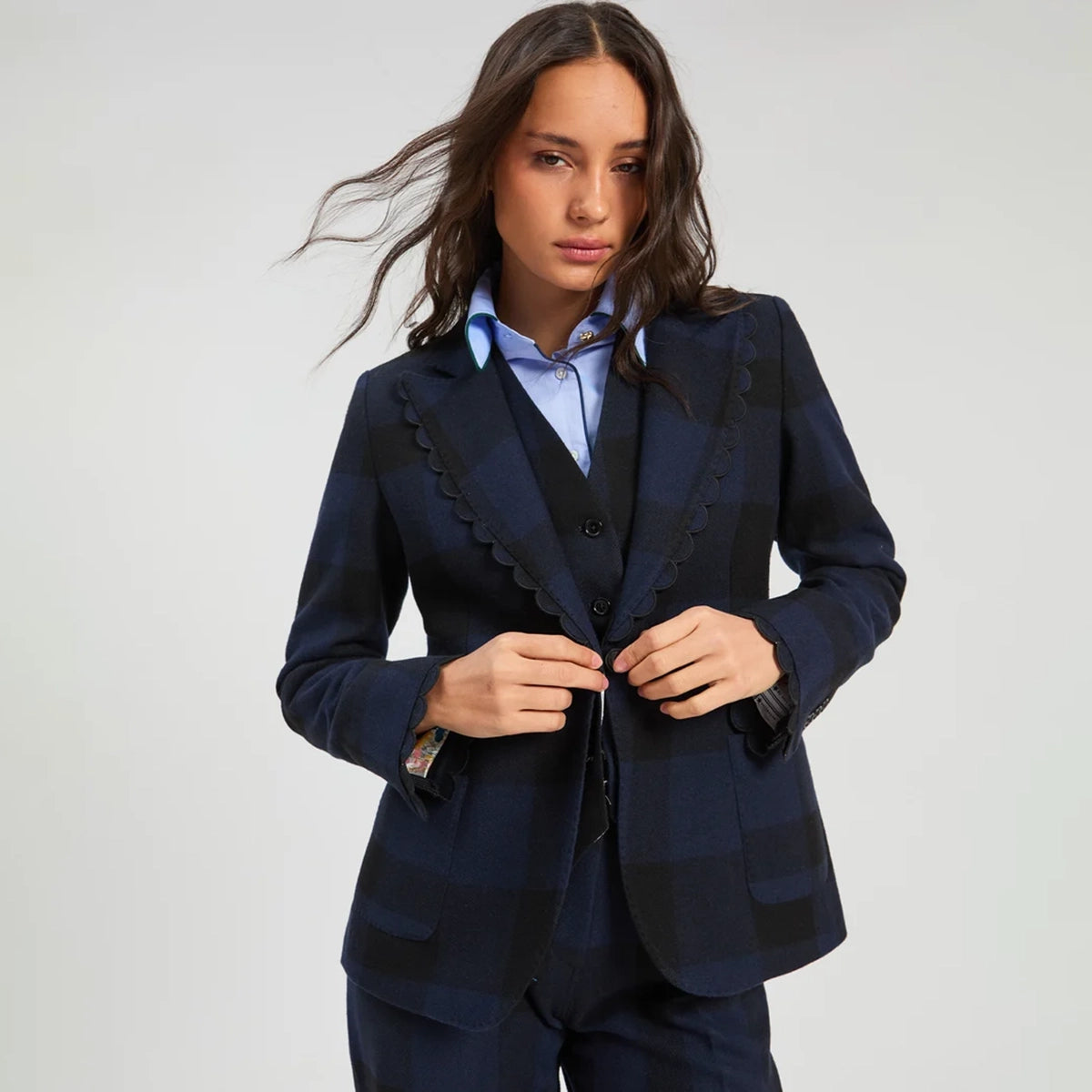 A woman modeling a dark navy blue plaid blazer with distinctive scalloped edges, worn over a matching vest and a light blue collared shirt, against a plain light gray studio background.