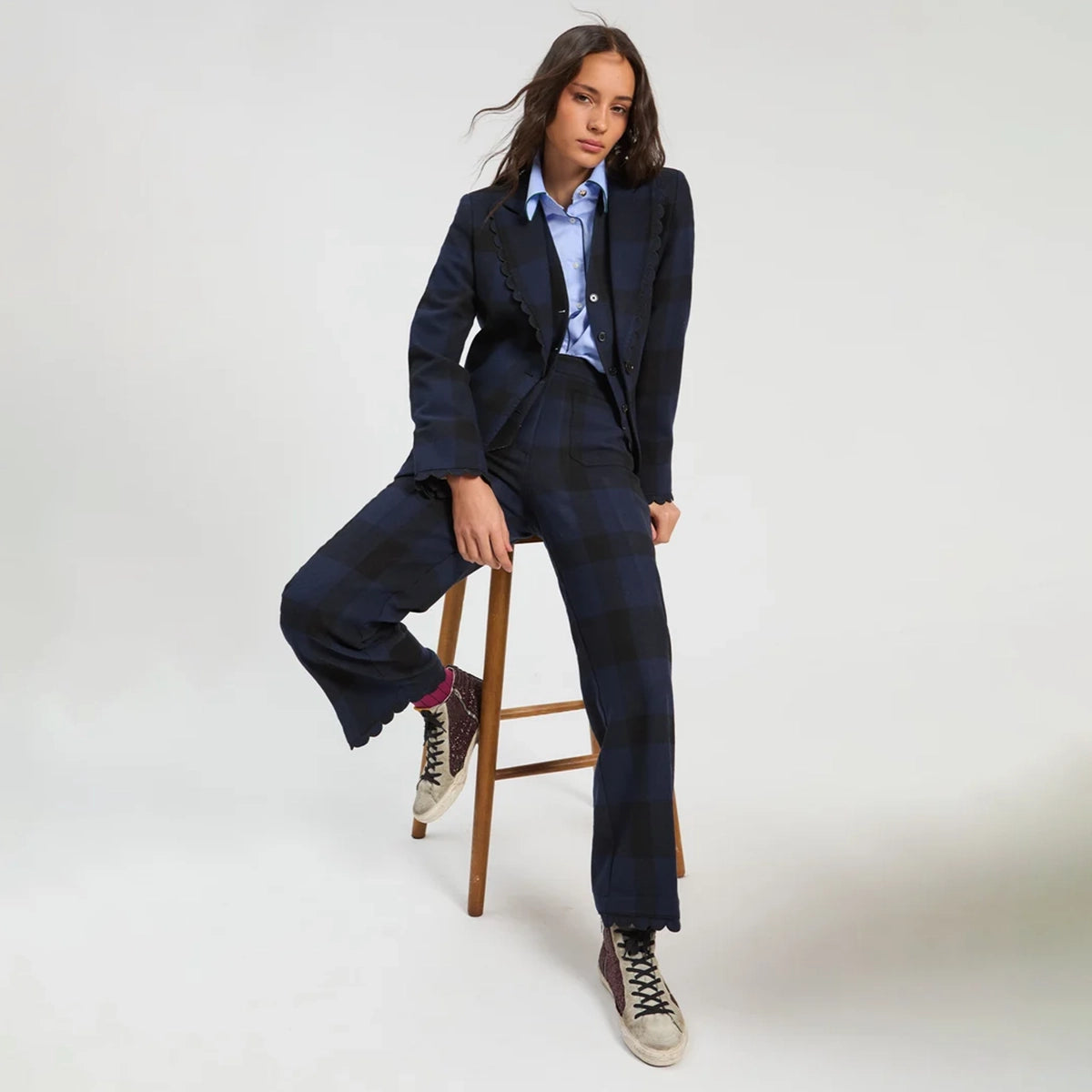 A model wearing a dark navy blue plaid blazer and matching wide-leg trousers, styled with a light blue button-down shirt, pink socks, and beige sneakers, sitting on a wooden stool against a light gray studio background.