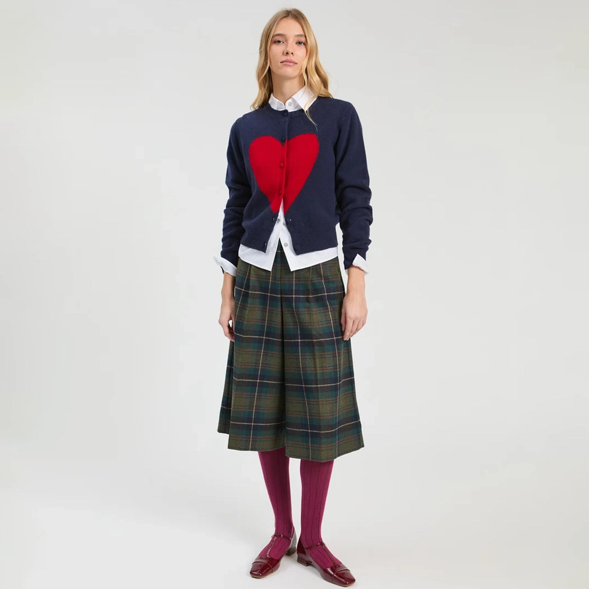 A model stands against a light gray studio background wearing a navy cardigan with a large red heart, a green plaid skirt, a white collared shirt, burgundy tights, and burgundy shoes.