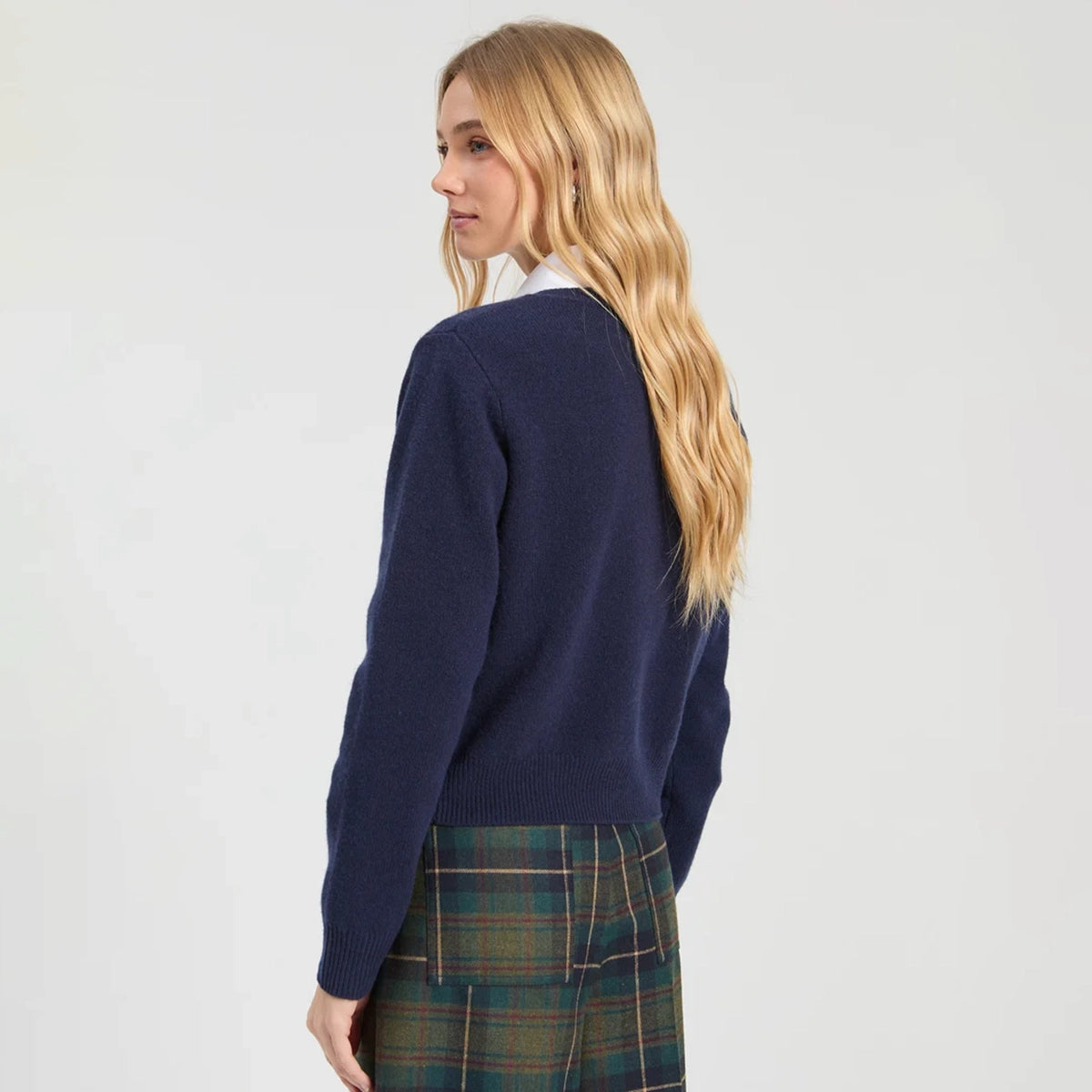 A woman with long blonde hair is shown from a side-back angle wearing a navy blue knit cardigan over a white collared shirt and a green plaid skirt, against a plain light gray studio background.