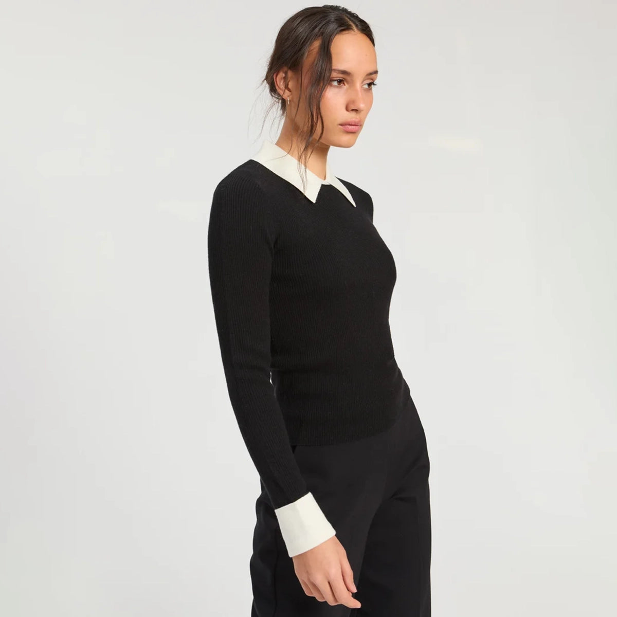 Model wearing a fitted black ribbed long-sleeve top with a contrasting cream pointed collar and matching cream cuffs, paired with black trousers.