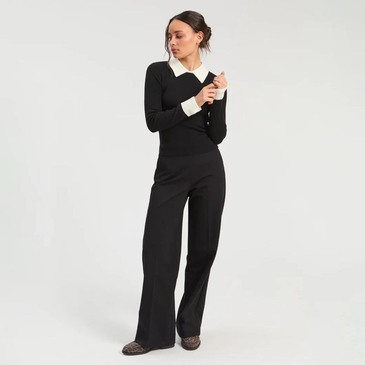Model wearing a fitted black ribbed long-sleeve top with a contrasting cream pointed collar and cuffs, paired with black wide-leg trousers and patterned flat shoes.