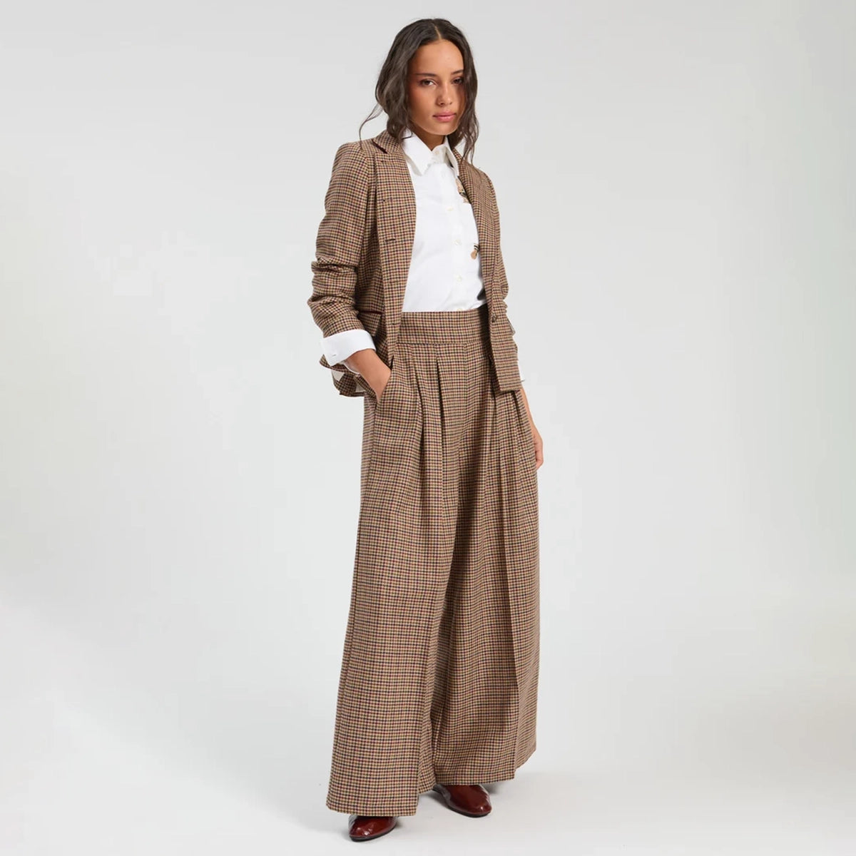 A woman models a brown, blue and green plaid wide-leg suit set, consisting of a blazer and matching pleated trousers, styled with a white collared shirt and brown shoes against a light gray studio background.