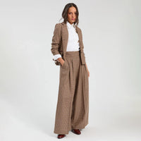 A woman models a brown, blue and green plaid wide-leg suit set, consisting of a blazer and matching pleated trousers, styled with a white collared shirt and brown shoes against a light gray studio background.