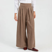 A model wearing high-waisted, wide-leg pleated trousers in a brown, blue and green plaid pattern, styled with a white long-sleeve shirt and dark brown leather loafers against a plain white studio background.