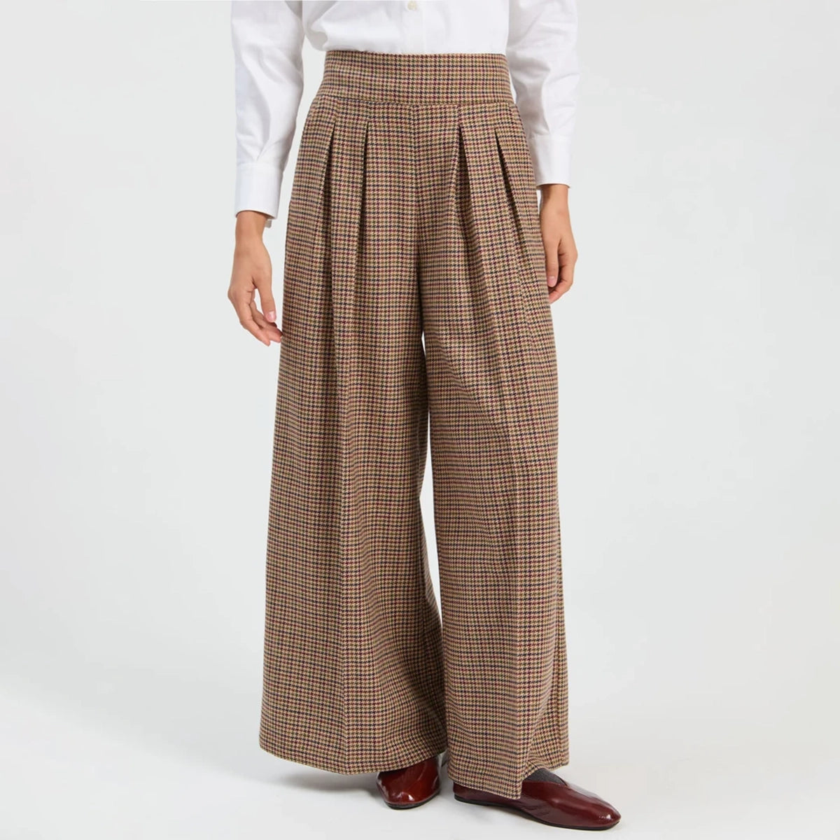 A model wearing high-waisted, wide-leg pleated trousers in a brown, blue and green plaid pattern, styled with a white long-sleeve shirt and dark brown leather loafers against a plain white studio background.