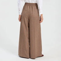 A person shown from the back wearing high-waisted, wide-leg trousers in a brown, blue and green plaid pattern, paired with a white long-sleeve blouse and dark flat shoes, against a plain white studio background.