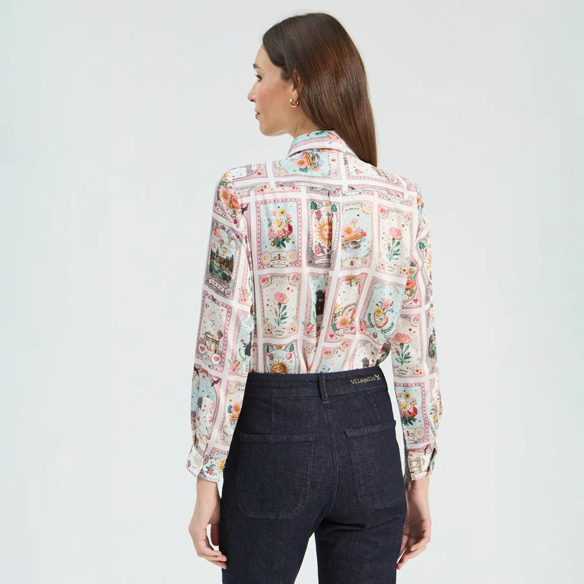 A woman shown from the back wearing a long-sleeve button-up shirt with a colorful vintage playing card pattern and dark navy jeans, set against a plain light gray studio background.