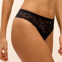 Black lace thong underwear with a sheer mesh panel, shown on a model against a light beige background.