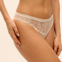 A close-up of a model wearing a pair of beige lace and mesh panties against a solid light beige background.