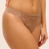 A close-up of a model wearing a beige lace thong with a floral lace pattern and a small rhinestone accent on the front, set against a plain light beige background.