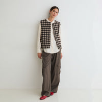 A model stands against a plain white studio wall wearing a black and beige plaid vest over a white button-up shirt, paired with wide-leg brown trousers and red shoes.