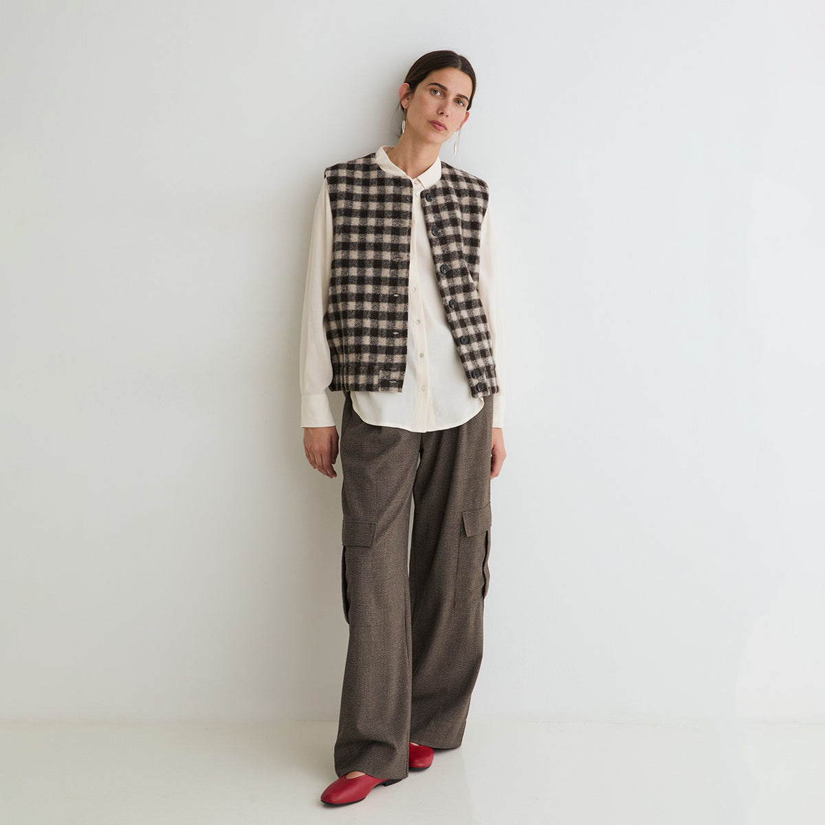 A model stands against a plain white studio wall wearing a black and beige plaid vest over a white button-up shirt, paired with wide-leg brown trousers and red shoes.