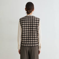 A model shown from the back wearing a sleeveless black and beige checkered vest over a cream long-sleeve blouse and brown trousers, set against a plain light gray background.